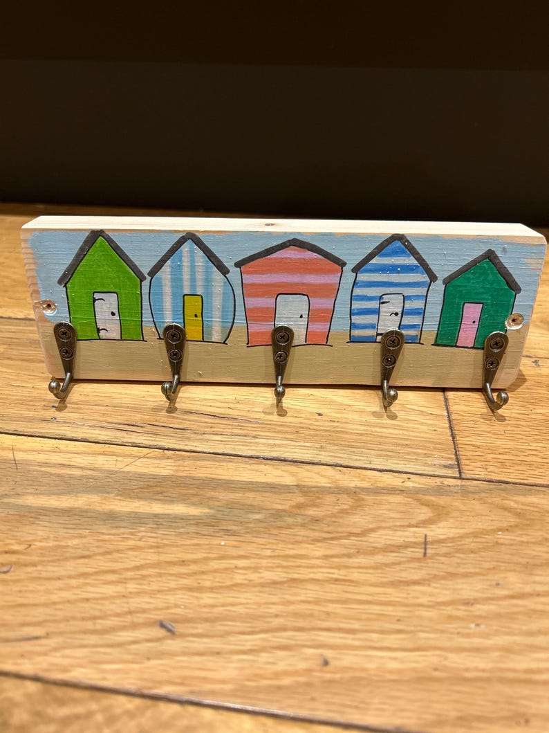 May include: A rectangular wooden wall hook rack featuring a hand-painted scene of five colourful beach huts in green, blue, and orange stripes. The piece includes five dark metal hooks attached with screws, set against a light blue sky and tan sand.