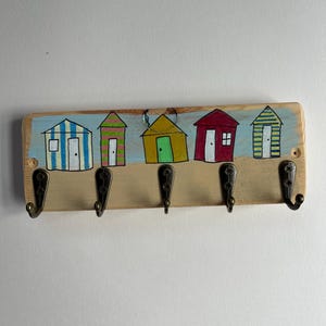 May include: A rectangular wooden wall hook rack featuring five dark metal hooks. The board is painted with a light blue sky and tan sand background, displaying five colourful beach huts in blue stripes, pink stripes, solid yellow, solid red, and yellow stripes.