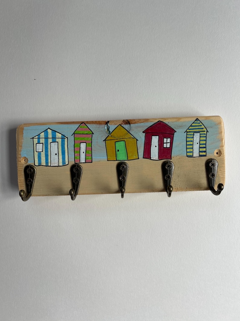 May include: A rectangular wooden wall hook rack featuring five dark metal hooks. The board is painted with a light blue sky and tan sand background, displaying five colourful beach huts in blue stripes, pink stripes, solid yellow, solid red, and yellow stripes.