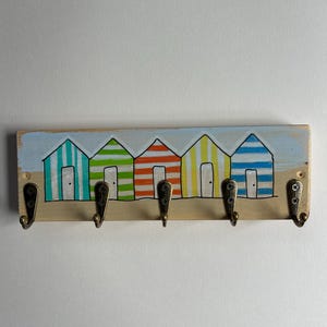 May include: A rectangular wooden wall hook rack featuring a hand-painted scene of five colourful striped beach huts in teal, green, orange, yellow, and blue. The rack includes five antique brass-toned metal hooks attached with visible screws.