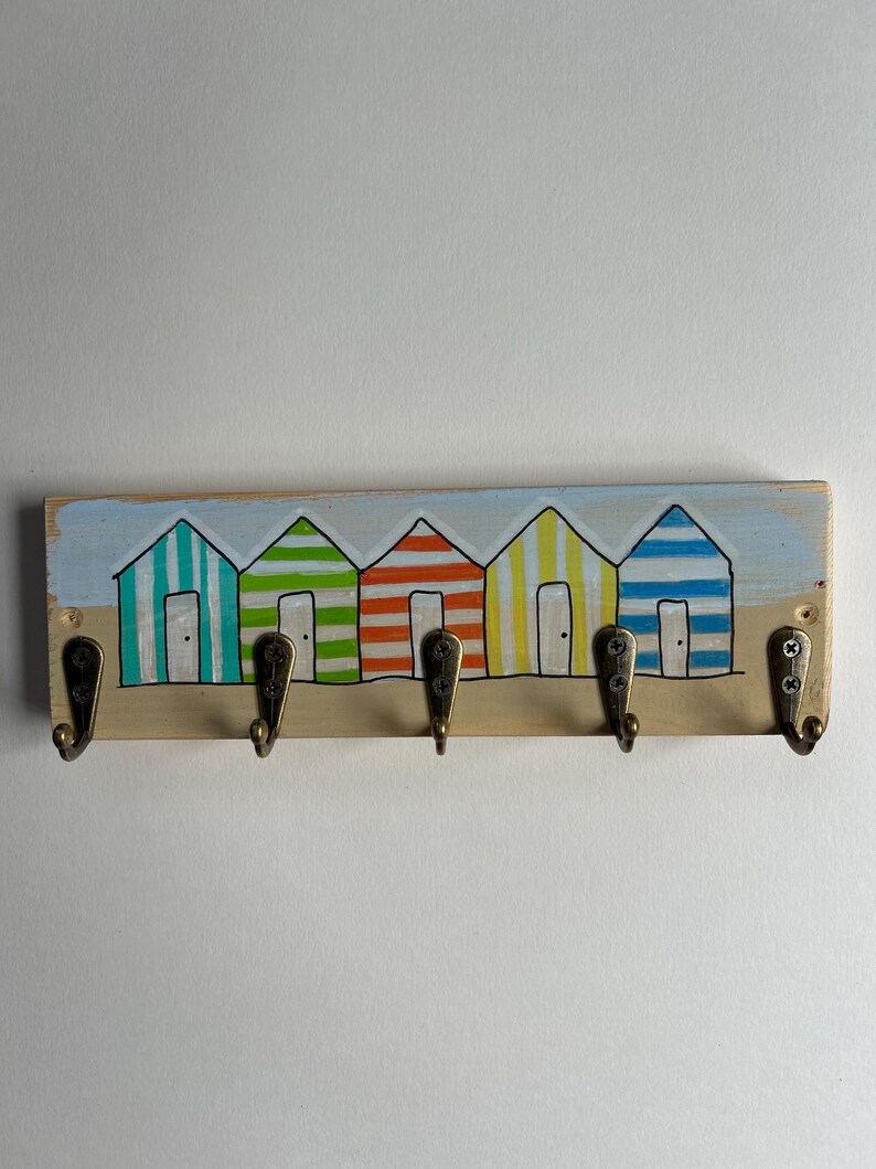 May include: A rectangular wooden wall hook rack featuring a hand-painted scene of five colourful striped beach huts in teal, green, orange, yellow, and blue. The rack includes five antique brass-toned metal hooks attached with visible screws.