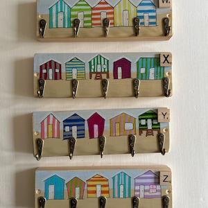 May include: Four rectangular wooden wall-mounted key racks, each featuring five bronze-coloured metal hooks. Each rack displays a hand-painted scene of five colourful beach huts on a sandy shore. Each rack includes a small wooden tile labelled W, X, Y, or Z.