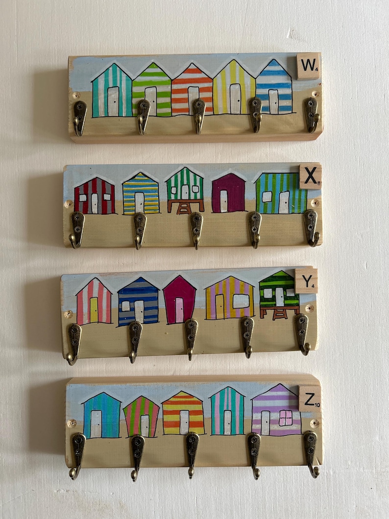 May include: Four rectangular wooden wall-mounted key racks, each featuring five bronze-coloured metal hooks. Each rack displays a hand-painted scene of five colourful beach huts on a sandy shore. Each rack includes a small wooden tile labelled W, X, Y, or Z.