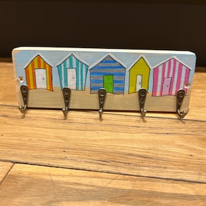 May include: A rectangular wooden wall hook rack featuring a hand-painted scene of five colourful beach huts in orange, teal, blue, yellow, and pink stripes. The base is painted with a light blue sky and tan sand, with five metal hooks.
