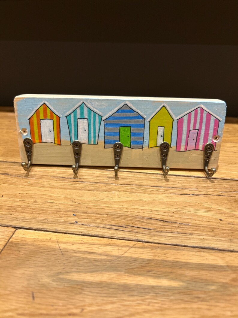 May include: A rectangular wooden wall hook rack featuring a hand-painted scene of five colourful beach huts in orange, teal, blue, yellow, and pink stripes. The base is painted with a light blue sky and tan sand, with five metal hooks.