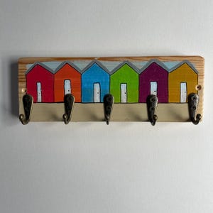 May include: A rectangular wooden wall hook rack featuring a painted row of six colourful beach huts in red, orange, blue, green, purple, and yellow. The rack includes five dark metal hooks attached to the bottom edge for hanging items.
