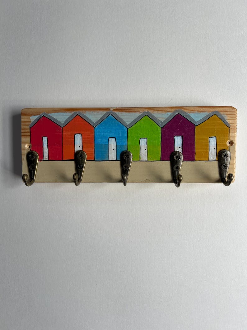 May include: A rectangular wooden wall hook rack featuring a painted row of six colourful beach huts in red, orange, blue, green, purple, and yellow. The rack includes five dark metal hooks attached to the bottom edge for hanging items.