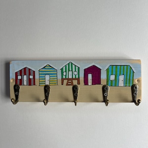May include: A rectangular wooden wall hook rack featuring a hand-painted scene of five colourful beach huts in red, yellow, blue, green, and purple. The rack includes five antique brass-toned metal hooks attached with screws to the lower section.