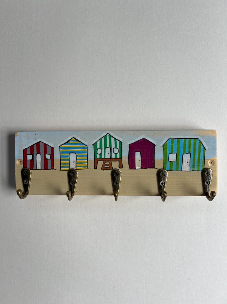 May include: A rectangular wooden wall hook rack featuring a hand-painted scene of five colourful beach huts in red, yellow, blue, green, and purple. The rack includes five antique brass-toned metal hooks attached with screws to the lower section.