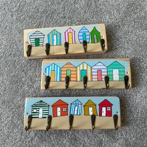May include: Three rectangular wooden wall hooks featuring hand-painted beach huts in various colours and patterns. Each piece includes five metal hooks attached to the bottom edge, displaying a light blue sky and sandy ground painted background.