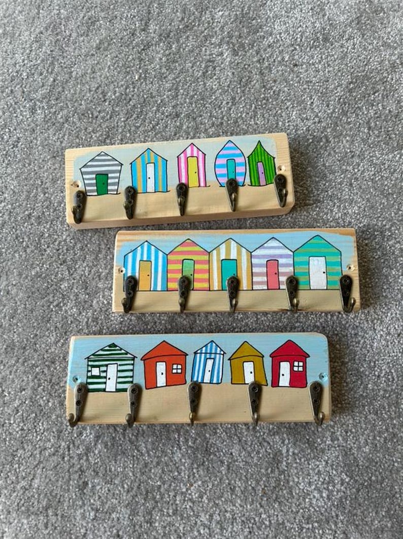May include: Three rectangular wooden wall hooks featuring hand-painted beach huts in various colours and patterns. Each piece includes five metal hooks attached to the bottom edge, displaying a light blue sky and sandy ground painted background.