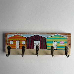 May include: A rectangular wooden wall hook rack featuring a hand-painted scene of three beach huts in orange, magenta, and teal stripes. The rack includes five antique brass-coloured metal hooks attached with screws along the bottom edge.