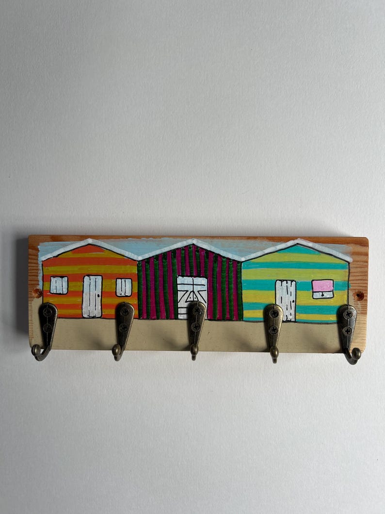 May include: A rectangular wooden wall hook rack featuring a hand-painted scene of three beach huts in orange, magenta, and teal stripes. The rack includes five antique brass-coloured metal hooks attached with screws along the bottom edge.