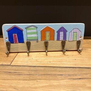 May include: A rectangular wooden wall hook rack featuring a hand-painted scene of five colourful beach huts in blue, green, orange, purple, and pink against a light blue sky. Five antique brass-toned metal hooks are attached to the lower section.