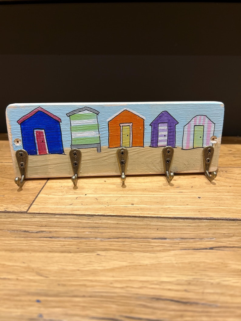 May include: A rectangular wooden wall hook rack featuring a hand-painted scene of five colourful beach huts in blue, green, orange, purple, and pink against a light blue sky. Five antique brass-toned metal hooks are attached to the lower section.