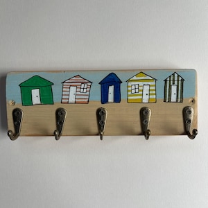 May include: A rectangular wooden wall hook rack featuring five metal hooks. The top section is painted with five colourful beach huts in green, orange-striped, blue, yellow-striped, and green-striped patterns against a light blue sky and sand.