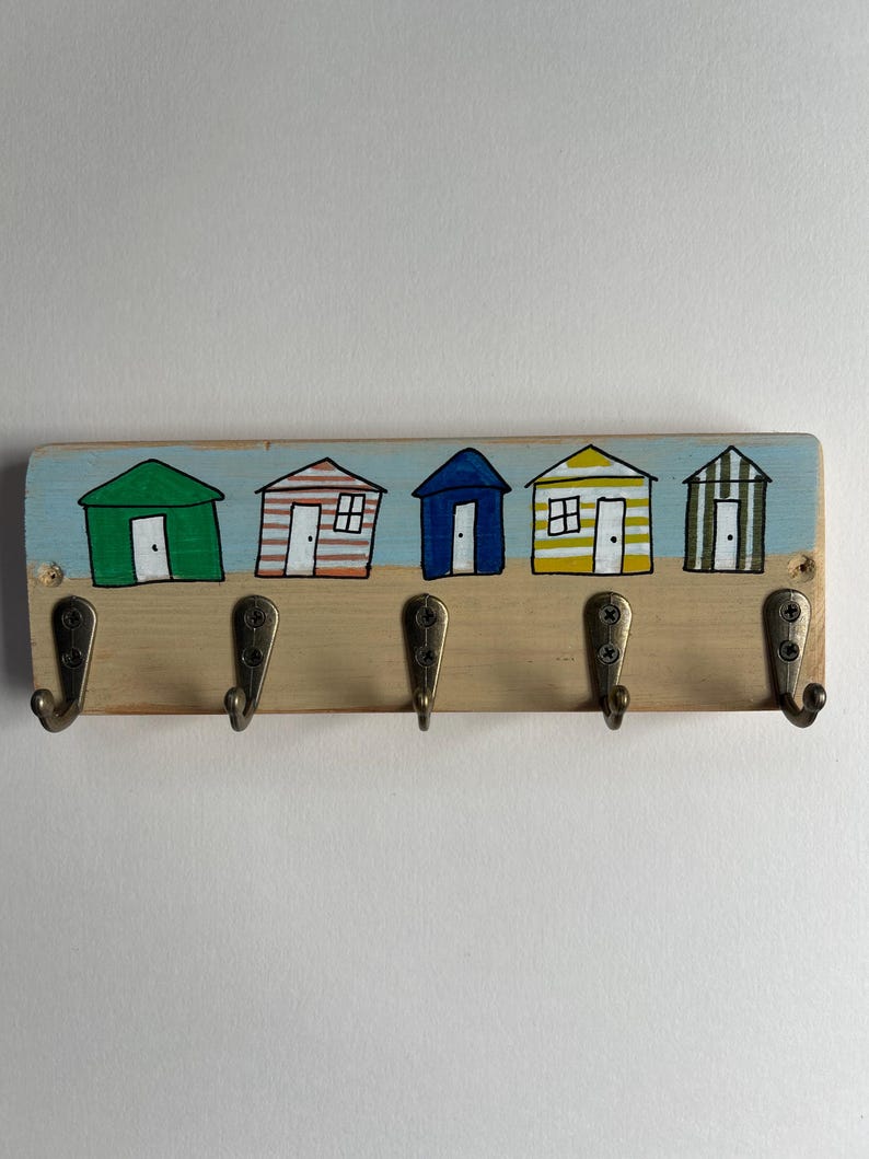 May include: A rectangular wooden wall hook rack featuring five metal hooks. The top section is painted with five colourful beach huts in green, orange-striped, blue, yellow-striped, and green-striped patterns against a light blue sky and sand.