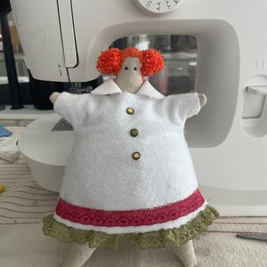 May include: A handmade doll with orange yarn hair, wearing a white dress with a red and green trim. The doll has a button closure and is positioned in front of a Singer sewing machine.