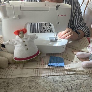 May include: A white Singer sewing machine is in use, with a small doll in a white coat and a stuffed animal on either side. The doll has red hair and a red trim on the coat. The stuffed animals are light pink and beige.
