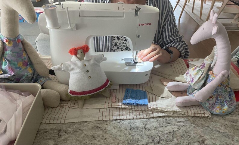 May include: A white Singer sewing machine is in use, with a small doll in a white coat and a stuffed animal on either side. The doll has red hair and a red trim on the coat. The stuffed animals are light pink and beige.