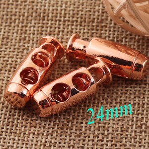 May include: Three rose gold toggle cord stoppers, each with two holes and a spring mechanism. The stoppers are 24 mm long.