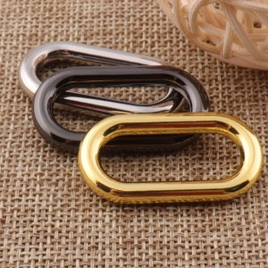 May include: Three oval-shaped metal rings in silver, black, and gold. The rings are stacked on top of each other, with the gold ring on the bottom.