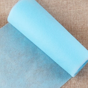 May include: A roll of light blue non-woven fabric. The fabric is unrolled and folded over on itself, showing the texture of the material.