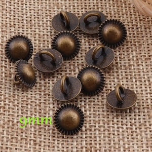 Puede incluir: Un conjunto de 12 botones de metal de color dorado antiguo con un borde festoneado decorativo y un pequeño lazo en la parte superior. Los botones miden aproximadamente 9 mm de diámetro.