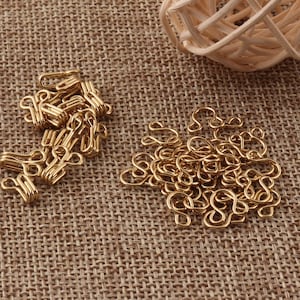 May include: A pile of gold-colored metal hook and eye closures. The hooks and eyes are arranged in a random pattern on a brown burlap surface.