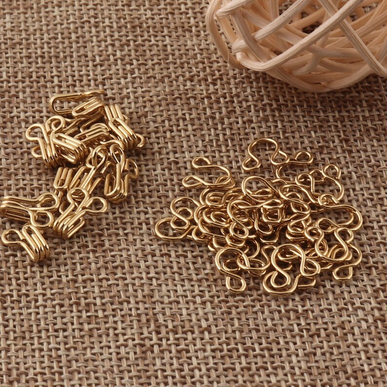 May include: A pile of gold-colored metal hook and eye closures. The hooks and eyes are arranged in a random pattern on a brown burlap surface.