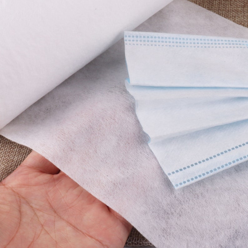 May include: A roll of white non-woven fabric and a folded blue surgical mask. The fabric is soft and textured. The mask has a blue and white pattern.