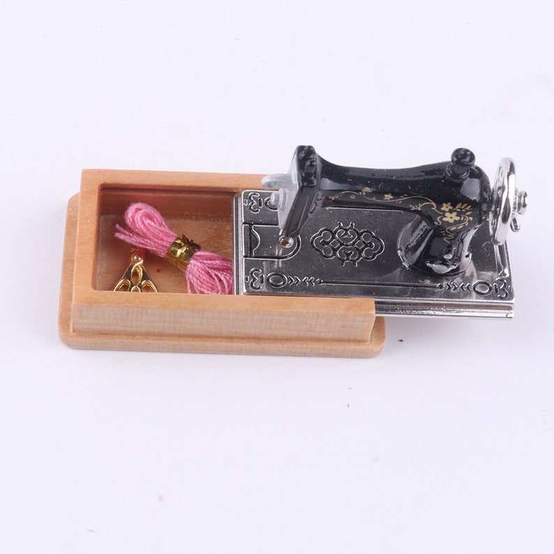 Może przedstawiać: Miniaturowa czarna maszyna do szycia z ozdobnym wzorem kwiatowym, otwarta, aby ujawnić szpulkę r&oacute;żowej nici i złotą szpulkę w drewnianym pudełku.