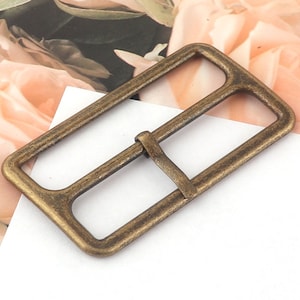 May include: A gold-toned metal buckle with a rectangular shape and a smooth, slightly textured surface.