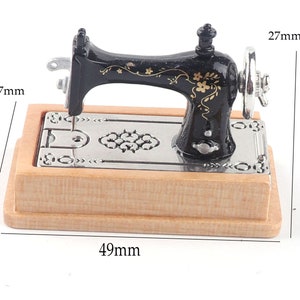 Może przedstawiać: Miniaturowa czarna maszyna do szycia z złotymi kwiatowymi detalami na drewnianej podstawie. Maszyna ma 27 mm wysokości i 49 mm szerokości.