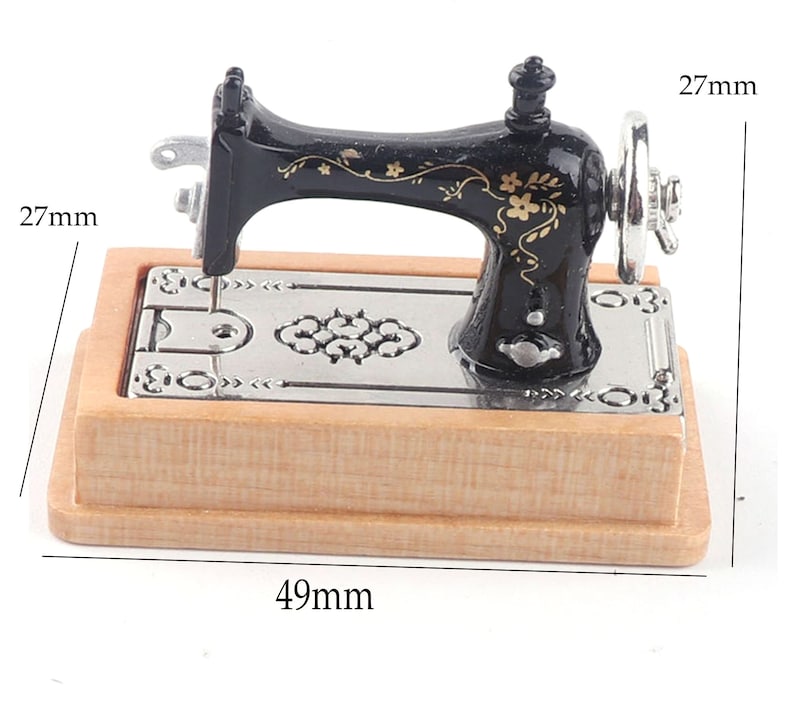 Może przedstawiać: Miniaturowa czarna maszyna do szycia z złotymi kwiatowymi detalami na drewnianej podstawie. Maszyna ma 27 mm wysokości i 49 mm szerokości.