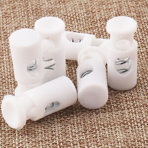 May include: Six white plastic cord locks with a metal spring inside. The cord locks are used to secure cords or ropes.