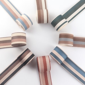 Puede incluir: Surtido de rollos de cinta a rayas en varias combinaciones de colores, incluyendo azul, rosa, marrón y verde. Las cintas están dispuestas en un patrón circular, mostrando sus diferentes patrones de rayas y colores. Las cintas están hechas de material tejido.