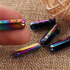 May include: A collection of iridescent metal shoelace aglets. The aglets are a mix of rainbow colors, including blues, greens, and yellows. Each aglet has a rectangular shape with a rounded end and small holes for the laces.