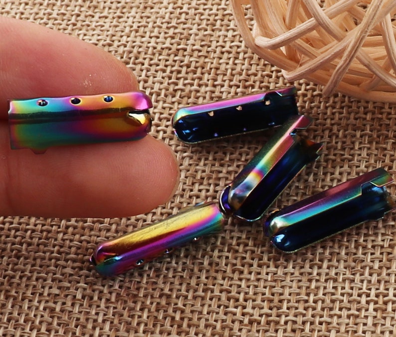 May include: A collection of iridescent metal shoelace aglets. The aglets are a mix of rainbow colors, including blues, greens, and yellows. Each aglet has a rectangular shape with a rounded end and small holes for the laces.