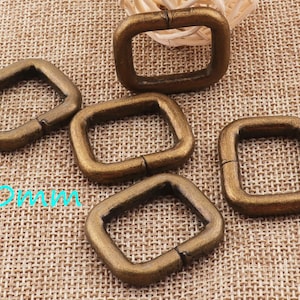 May include: Five gold-toned metal D-rings, each measuring 20mm in diameter. The rings are arranged in a semi-circle on a brown burlap fabric background.