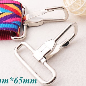 Könnte beinhalten: Zwei silberne Metallschließen mit einem Haken und einem D-Ring. Die Schließen sind 50 mm x 65 mm groß.