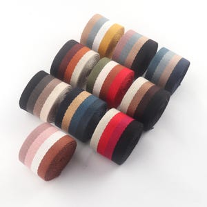 Op de afbeelding: Een verzameling kleurrijke gestreepte lintrollen. Elke rol heeft een unieke combinatie van kleuren, waaronder zwart, bruin, rood, blauw en wit. De linten zijn strak opgerold en worden tegen een witte achtergrond getoond.