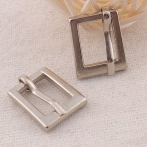 May include: Two silver metal belt buckle slides. The slides are rectangular with a rounded edge and a single hole on the top. The slides are designed to be used with a belt strap.