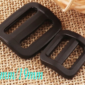May include: Two black plastic slider buckles, one measuring 13mm and the other 19mm. The buckles are on a woven brown fabric background.