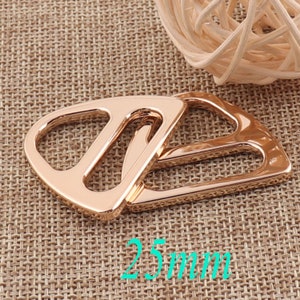 Pode incluir: Duas fivelas deslizantes de metal dourado, cada uma com forma triangular arredondada e uma abertura central. As fivelas medem 25 mm.