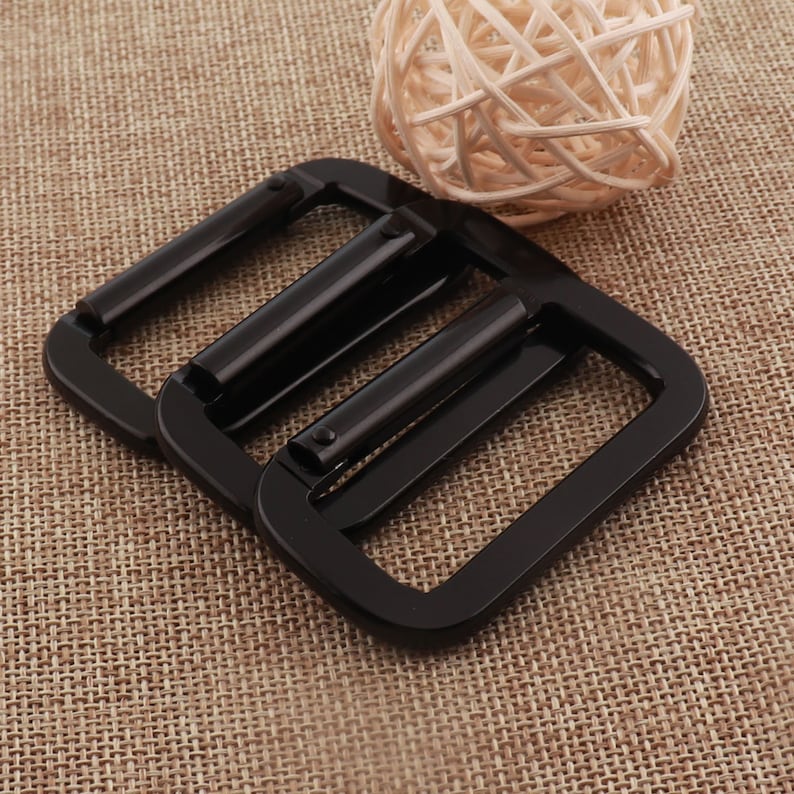 May include: Two black metal belt buckle slides. The slides are rectangular with rounded corners and have a smooth, shiny finish.