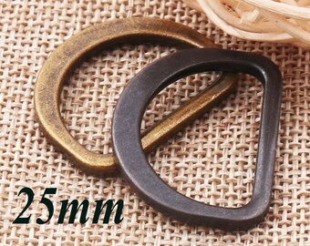 Puede incluir: Dos anillos en D de metal, uno dorado y otro negro, sobre un fondo de tejido marrón tejido. Los anillos tienen un diámetro de 25 mm.