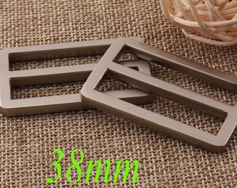 May include: Two silver metal slider buckles, each with two parallel slots, measuring 38 mm in length. The buckles are on a brown woven fabric background.
