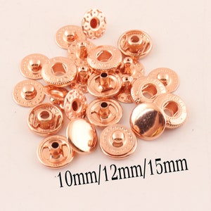 Könnte beinhalten: Eine Sammlung von 10 mm, 12 mm und 15 mm großen Druckknöpfen aus roségoldfarbenem Metall. Die Druckknöpfe sind rund und haben eine glatte, glänzende Oberfläche.