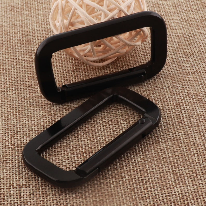 May include: Two black metal D-shaped carabiners, one on top of the other, on a brown fabric background.